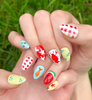 Two hands displaying summer press on nails with vibrant, fun designs. Nail designs include a lobster, stars, cherries, gingham, fish, watermelon, and blueberries. Nail colors include red, white, orange, light blue, green, and yellow. The two hands are displaying the press on nails on a grassy summer background.