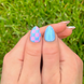 Colorful press-on nail art on thumbs. One thumb has pink and blue checkered design and light blue with pink and white dots on the other thumb. Summer Nails