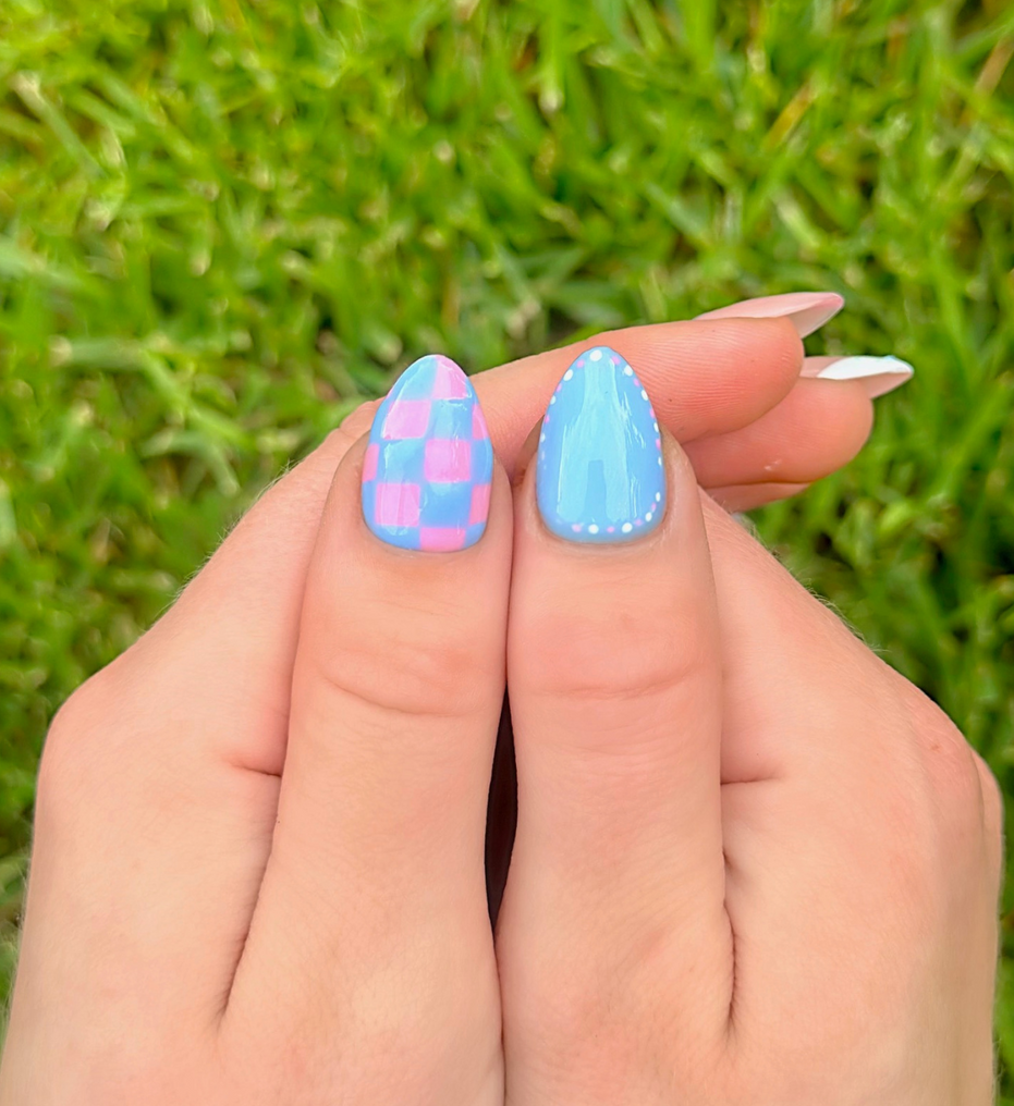 Colorful press-on nail art on thumbs. One thumb has pink and blue checkered design and light blue with pink and white dots on the other thumb. Summer Nails