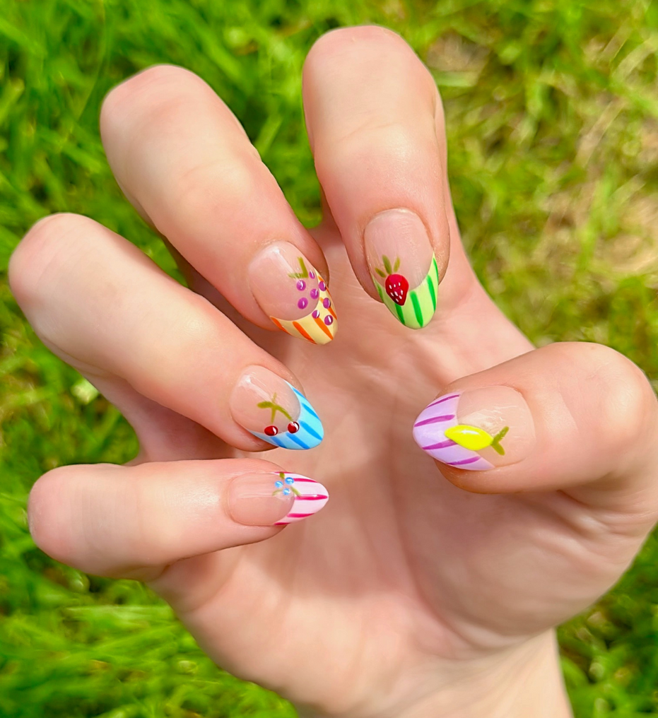 One hand showing Fruity Press On Nails with vibrant colors.  The press ons have colorful french striped tips and different fruits on each nail including grapes, cherries, lemon, blueberries, and strawberries. Hands are stacked on top of each other to display each press on design.
