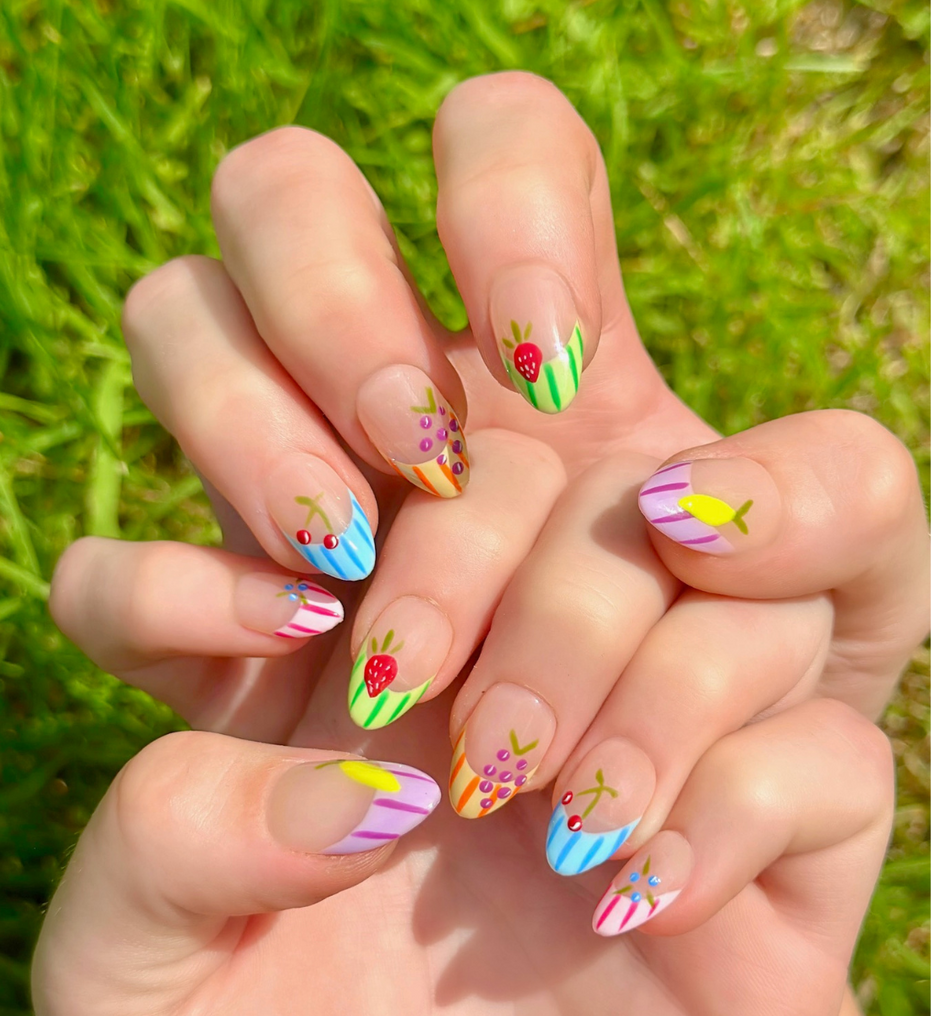 Two hands displaying Fruity Press On Nails with vibrant colors. Nails have colorful french striped tips and different fruits on each nail including grapes, cherries, lemon, blueberries, and strawberries. Hands are stacked on top of each other to display each press on design.
