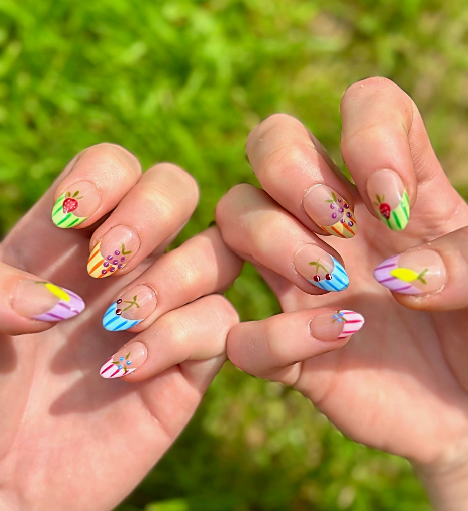 Two hands showing fruity Press On Nails with vibrant colors. Nails have colorful french striped tips and different fruits on each nail including grapes, cherries, lemon, blueberries, and strawberries. Hands are stacked on top of each other to display each press on design.