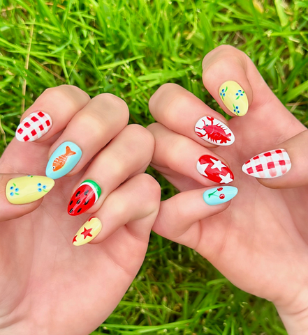 Two hands displaying summer press on nails with vibrant, fun designs. Nail designs include a lobster, stars, cherries, gingham, fish, watermelon, and blueberries. Nail colors include red, white, orange, light blue, green, and yellow. The two hands are displaying the press on nails on a grassy summer background.