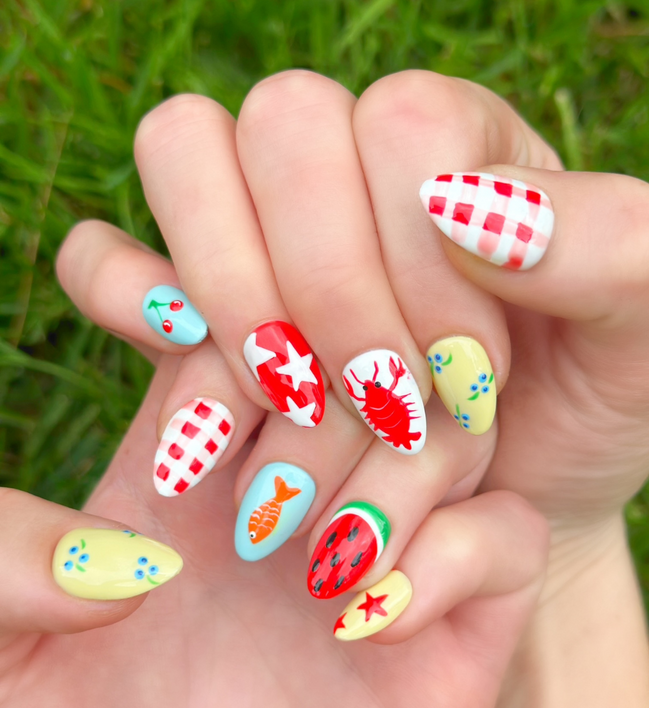 Two hands displaying summer press on nails with vibrant, fun designs. Nail designs include a lobster, stars, cherries, gingham, fish, watermelon, and blueberries. Nail colors include red, white, orange, light blue, green, and yellow. The two hands are displaying the press on nails on a grassy summer background.