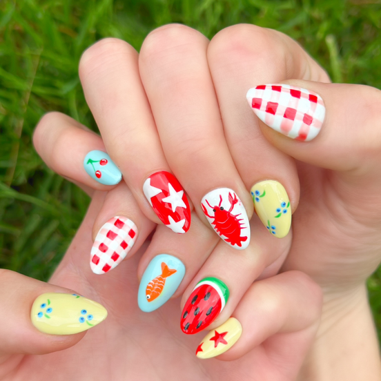 Two hands displaying summer press on nails with vibrant, fun designs. Nail designs include a lobster, stars, cherries, gingham, fish, watermelon, and blueberries. Nail colors include red, white, orange, light blue, green, and yellow. The two hands are displaying the press on nails on a grassy summer background.