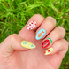 One hand displaying summer press on nails with vibrant, fun designs. Nail designs include a fish, stars,, gingham, watermelon, and blueberries. Nail colors include red, white, orange, light blue, green, and yellow. The two hands are displaying the press on nails on a grassy summer background.