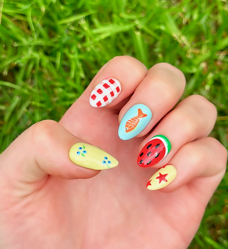One hand displaying summer press on nails with vibrant, fun designs. Nail designs include a fish, stars,, gingham, watermelon, and blueberries. Nail colors include red, white, orange, light blue, green, and yellow. The two hands are displaying the press on nails on a grassy summer background.