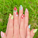 Two hands displaying summer press on nails with vibrant, fun designs. Nail designs include a lobster, stars, cherries, gingham, fish, watermelon, and blueberries. Nail colors include red, white, orange, light blue, green, and yellow. The two hands are displaying the press on nails on a grassy summer background.