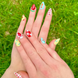 Two hands displaying summer press on nails with vibrant, fun designs. Nail designs include a lobster, stars, cherries, gingham, fish, watermelon, and blueberries. Nail colors include red, white, orange, light blue, green, and yellow. The two hands are displaying the press on nails on a grassy summer background.