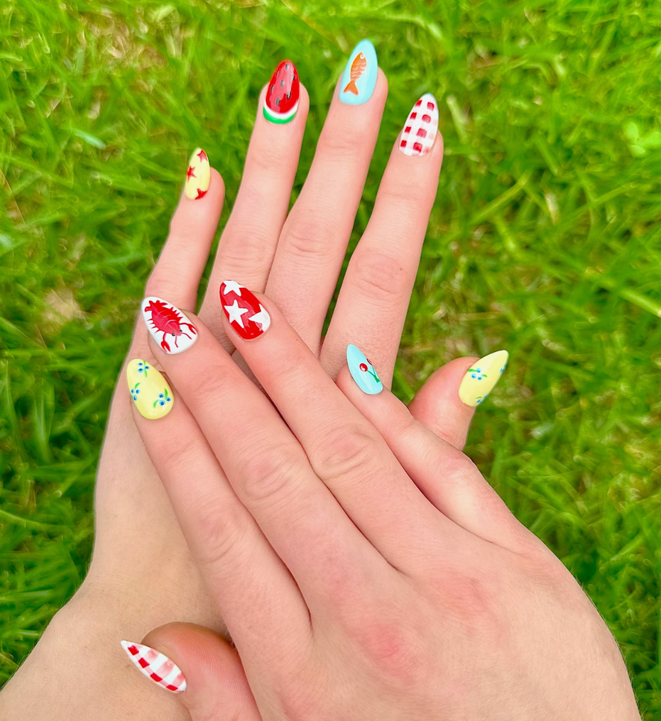 Two hands displaying summer press on nails with vibrant, fun designs. Nail designs include a lobster, stars, cherries, gingham, fish, watermelon, and blueberries. Nail colors include red, white, orange, light blue, green, and yellow. The two hands are displaying the press on nails on a grassy summer background.