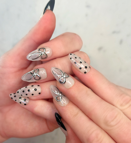 Hands with hand painted salon press-on nails in long almond shape, featuring black and silver polka dots, bows, and french tips