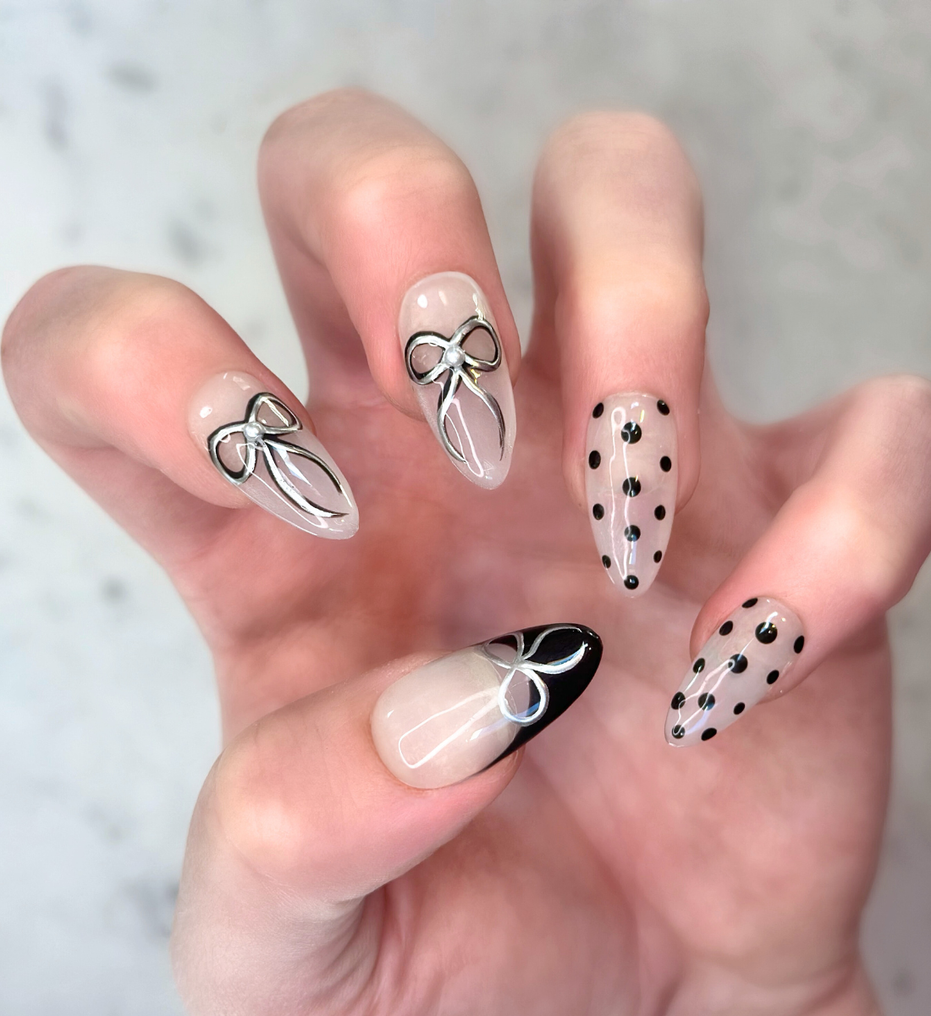 Hand with decorative nail art on salon press-ons featuring  black and silver bows, black polka dots, and black french tips with silver bow detail