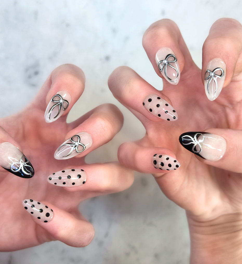 Two hands with hand painted salon press-on nails in long almond shape, featuring black and silver polka dots, bows, and french tips on neutral background