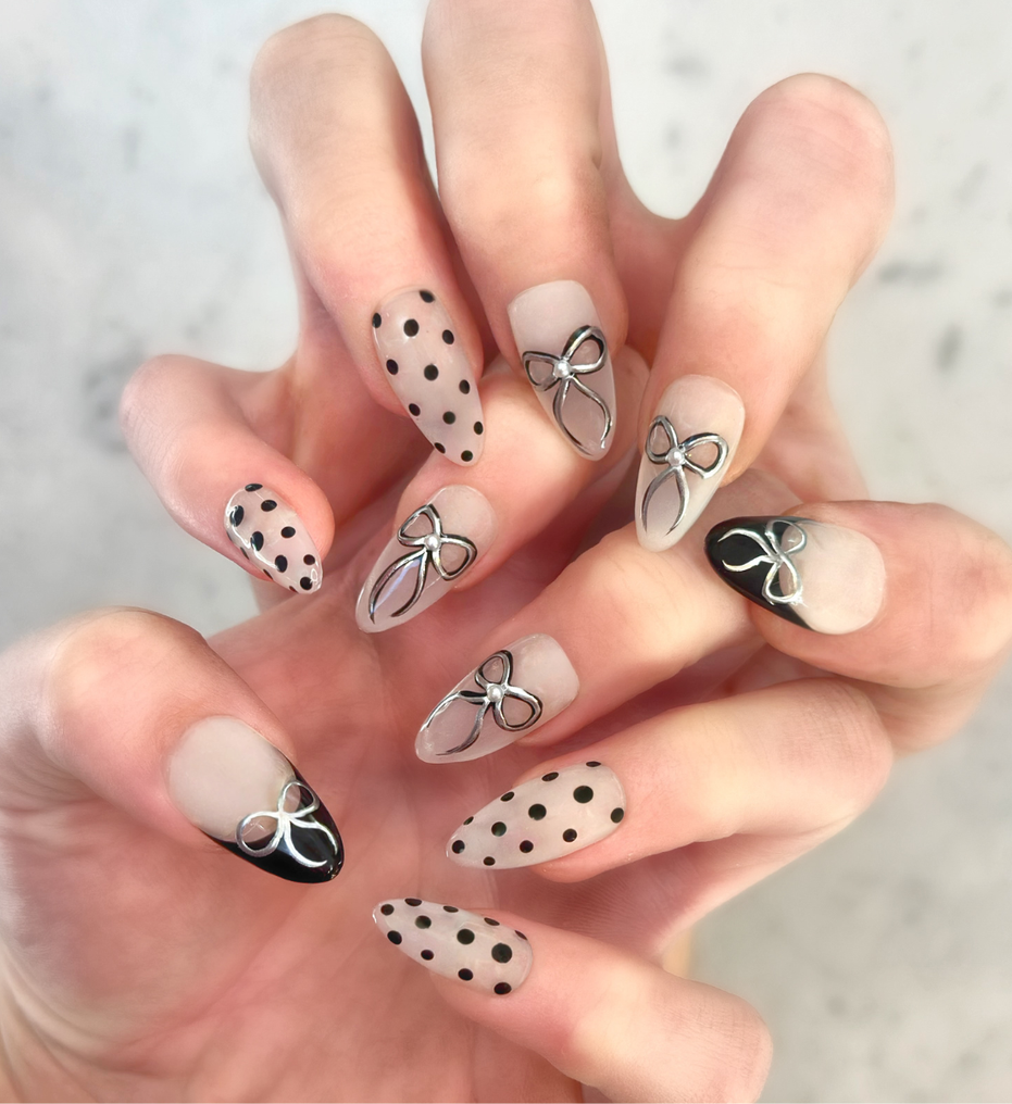 Two hands interlocked with hand painted salon press-on nails in long almond shape, featuring black and silver polka dots, bows, and french tips
