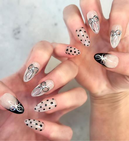 Two hands showing nails with hand painted salon press-on nails in long almond shape, featuring black and silver polka dots, bows, and french tips with silver bows on neutral background
