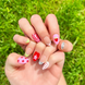Hand-Painted acrylic press-on nails with vibrant colors and designs including strawberries, stars, gingham, zebra print, and french tips. The nail colors are red, light green, light pink, zebra print, and silver details. Hands and nails are displayed on grassy summer background.