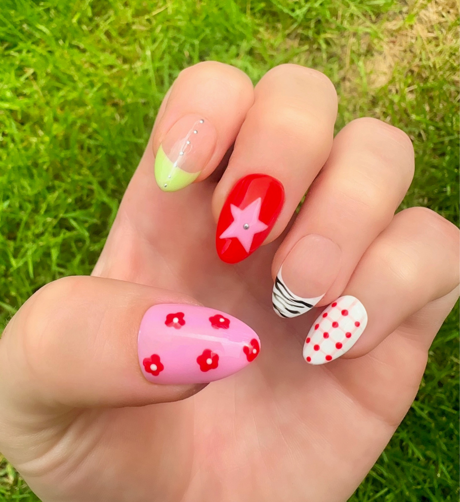Hand-Painted acrylic press-on nails with vibrant colors and designs including strawberries, stars, gingham, zebra print, and french tips. The nail colors are red, light green, light pink, zebra print, and silver details. The hand is displayed on a grass background.