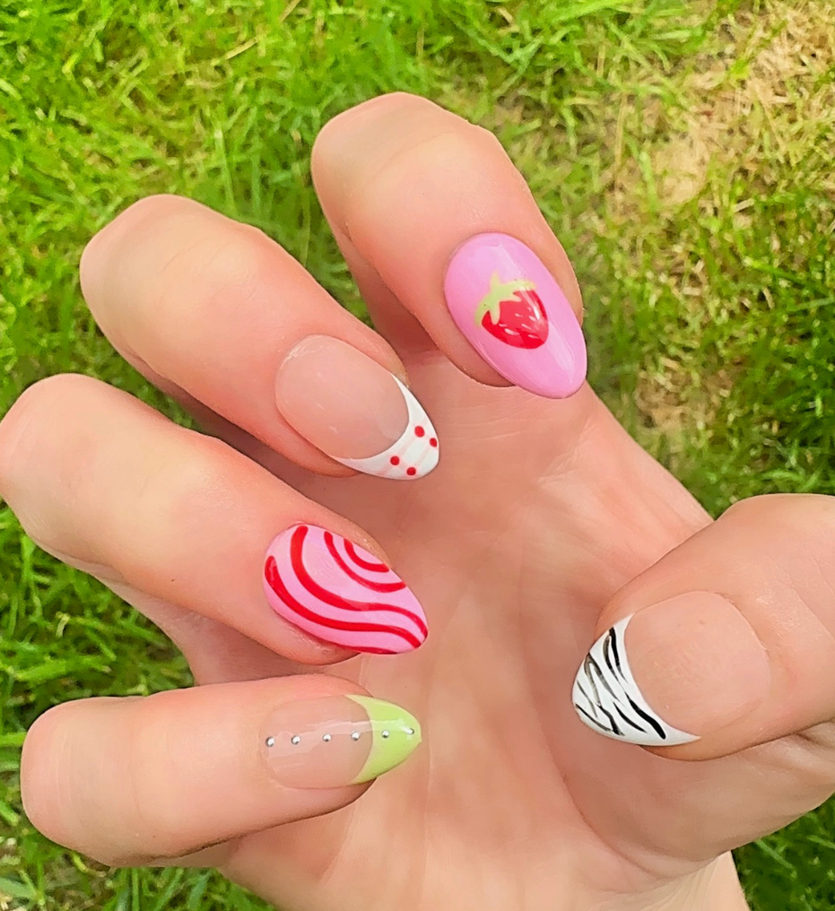 Hand-Painted acrylic press-on nails with vibrant colors and designs including strawberries, stars, gingham, zebra print, and french tips. The nail colors are red, light green, light pink, zebra print, and silver details. Two hands and nails are displayed on a grassy summer background.