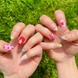 Hand-Painted acrylic press-on nails with vibrant colors and designs including strawberries, stars, gingham, zebra print, and french tips. The nail colors are red, light green, light pink, zebra print, and silver details. Two hands and nails are displayed on a grassy summer background.