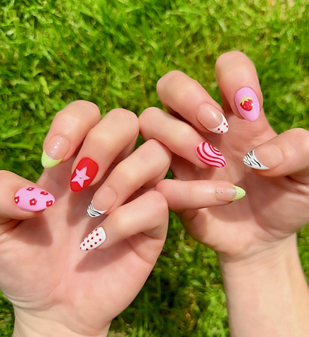 Hand-Painted acrylic press-on nails with vibrant colors and designs including strawberries, stars, gingham, zebra print, and french tips. The nail colors are red, light green, light pink, zebra print, and silver details. Two hands and nails are displayed on a grassy summer background.
